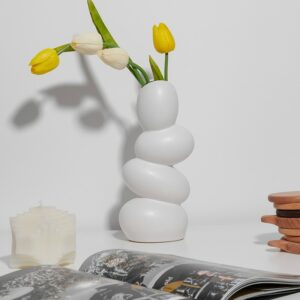Vase blanc moderne et original. Il est constitué d'alvéoles rondes, comme des œufs superposés. Sa silhouette déstructurée importe un charme novateur dans votre décoration d'intérieur. Le vase contient des fleurs tulipes jaunes et blanches. Le vase est exposé sur une surface blanche, à proximité d'un magazine, et d'une bougie.