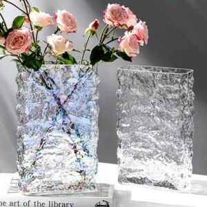 Vase en verre à la texture d'un glaçon. Ce vase glacé est rectangulaire, transparent et ondulé, avec les aspérités propres à la glace. L'un des deux vases s'éclaire de reflets multicolores, et contient un bouquet de rose. Il est exposé sur un livre blanc, sur une table blanche, devant un mur gris.