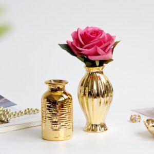 Vase céramique doré miniature. Deux vases sont exposés, l'un en forme de petite bouteille, l'autre en forme évasée, rappelant la silhouette d'un bonbon. Ce dernier contient une rose. Les deux vases sont brillants, avec des reflets lumineux. Ils sont exposés sur une surface blanche, a proximité de bijoux dorés et de magazines.