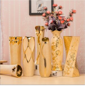 Vase vintage doré de forme tubulaire, présenté avec différentes finitions et gravures. C'est un vase résolument singulier qui se caractérise par son aspect rutilant et ses formes originales. Un des vases contient un bouquet de fleurs roses. Ces vases sont exposés dans un intérieure, sur une surface beige et devant un mur blanc et rose.