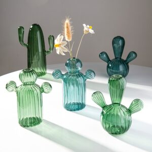 Vase original en forme de cactus transparent. Ce vase est confectionné en verre transparent, dans un design moderne, mignon et sympathique. Il est exposé en différentes formes de cactus. Il y a deux vases en cactus bleu et trois vases en cactus vert. Le vase central contient trois tiges de fleurs séchées de type pampa. Les vases sont posés sur une surface blanche.