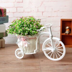 Vase rotin original en forme de petit tricycle blanc. Il contient un bouquet de fleurs artificielles dans un panier tressé entouré d'un ruban rose. Le vase est exposé à même le sol, sur un parquet avec du mobilier en arrière-plan et un mur en brique rose pâle.
