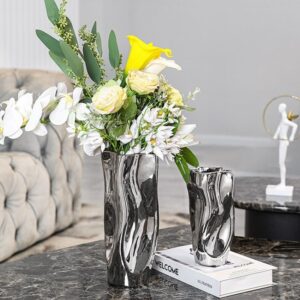 Vase céramique plissé de couleur argent. Le vase est exposé en duo, dans deux formats différents. Le plus grand vase contient un bouquet de fleurs blanches et jaunes, avec des tiges de plantes vertes. Le vase offre l'aspect du mercure, à la fois liquide et réfléchissant. Les vases sont exposés sur une table en marbre gris, avec un canapé en arrière-plan et une statuette blanche.