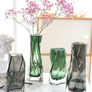 Vase en verre plissé, de forme géométrique, carré et torsadé. Les vases sont exposés en deux formats, petit et grand, ainsi qu'en deux couleurs, vert et gris. Le verre est teinté légèrement, mais offre un effet de transparence. Le grand vase vert contient un bouquet de petites fleurs roses. Ils sont exposés sur une table blanche, à proximité d'une statue grecque.