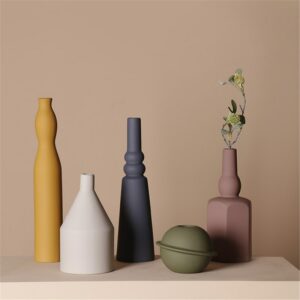 Vase scandinave moderne au design géométrique. Ce vase est exposé dans différente forme, de la boule à la silhouette tubulaire, et dans cinq coloris différents : Jaune, bleu, blanc , vert et rose. Le vase rose contient une tige de plante verte. Ils sont exposés sur une surface beige, devant un mur beige.