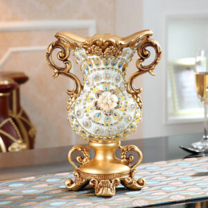 Grand vase de luxe prestigieux, de couleur nacre avec un pied et des anses dorées. Le vase est serti de brillants et intègre en son centre un motif floral. Il est exposé sur une table, dans un salon.