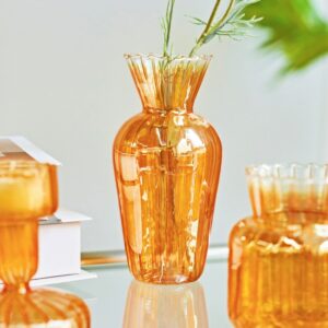 Vase en verre orange à l'apparence d'un bonbon acidulé. Ce vase brillant est disponible en différente forme. Il est exposé sur une table en verre à proximité d'un livre, et contient des tiges de plantes vertes.