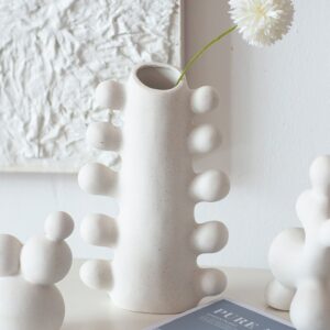 Vase céramique blanc dans un design moderne à l'apparence d'un cactus. Il est exposé en soliflore avec une fleur blanch à l'intérieur. Le vase est positionné sur une surface blanche, à proximité d'autres éléments décoratifs de mêmes couleurs, devant un mur blanc.