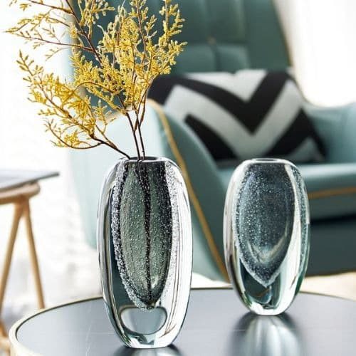 Deux vases de style Murano gris en verre à double paroi. Le coeur du vase est constitué de fines bulles. Il y a un vase petit format et un grand format qui contient une branche de mimosa. Ils sont disosés sur une table noire avec en fond du mobilier de salon composé d'un canapé vert avec un coussin blanc et noir.