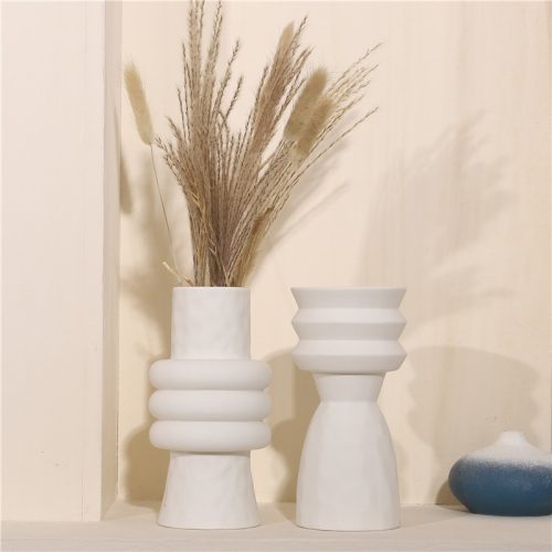 Vase scandinave blanc en céramique de style géométrique et moderne. Il est exposé en deux silhouettes différente, de forme évasée, tubulaire et arrondie. Un des vases contient un bouquet de fleurs de pampa. Ces vases sont exposés sur une étagère, devant un mur beige, rose.