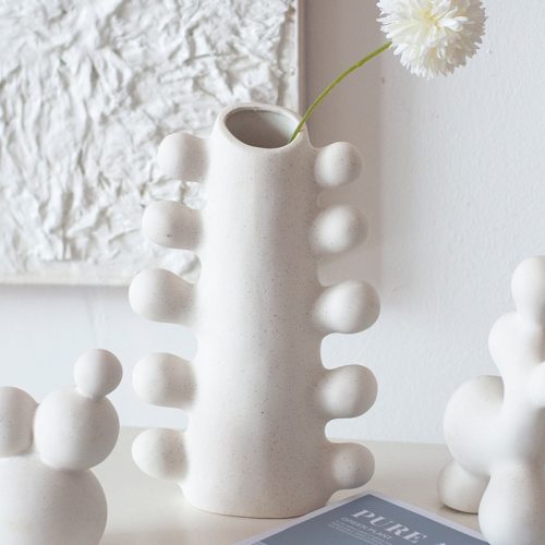 Vase céramique blanc dans un design moderne à l'apparence d'un cactus. Il est exposé en soliflore avec une fleur blanch à l'intérieur. Le vase est positionné sur une surface blanche, à proximité d'autres éléments décoratifs de mêmes couleurs, devant un mur blanc.
