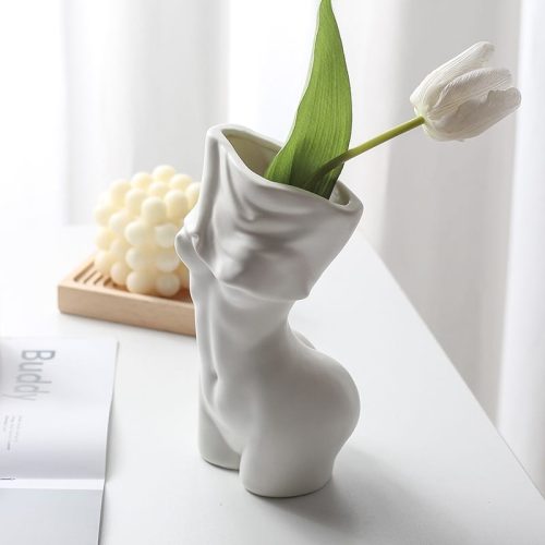 Vase blanc en forme de buste de femme nue. Celle-ci est en train de se déshabiller. Le vase est fait en céramique blanche. Il contient une tulipe blanche. Il est posé sur une table blanche avec un magazine ouvert à proximité, et une bougie en arrière-plan.
