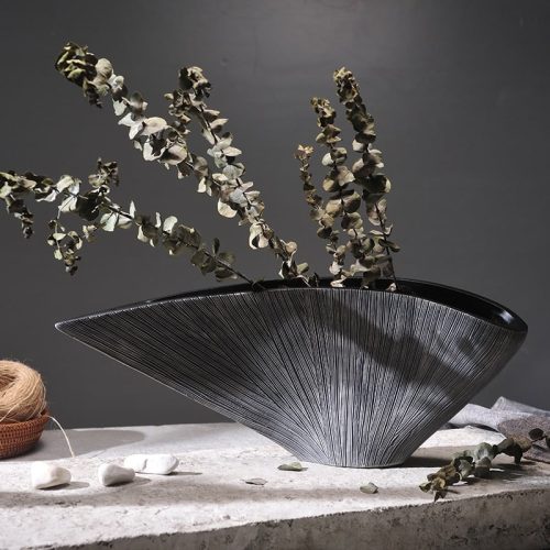 Grand vase en forme de coquillage noir. Inspiré du style japonais, il est très large et évasé. Sa céramique est brillante et finement striée. Il contient des branches de fleurs séchées. Il est entreposé sur une table en pierre avec du sable et des graviers.