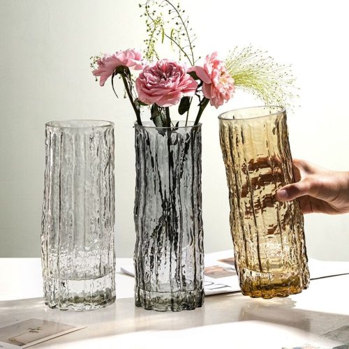 Trois vases à fleurs en verre, de forme tubulaire, avec une surface irrégulière plein de relief. Un vase est transparent, l'autre noir, et le dernier orange. Le noir contient des fleurs roses et quelques brins d'herbes.