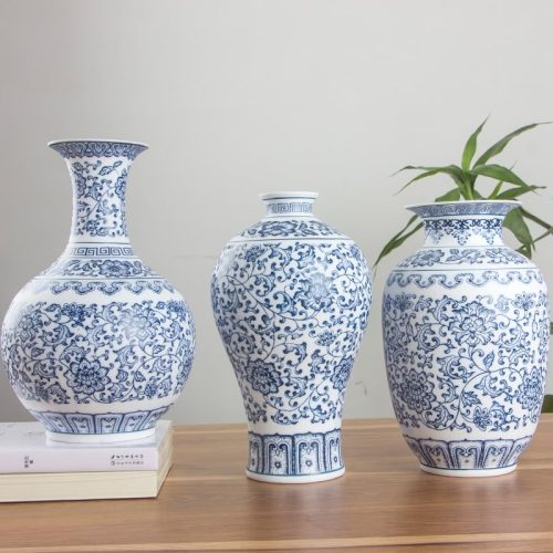 Trois vases en porcelaine blanc et bleu. Ils sont de style chinois, avec des motifs floraux et des arabesques typiques du style traditionnel. Ils ont trois formes différentes. L'un est arrondie à sa base et doté d'un col allongé avec une bouche évasée. Le deuxième à une forme d'urne, fine à la base et évasée en haut avec un col court. Le dernier à une forme ovale, plus classique, en sorte de petite jarre. Ils sont disposés sur une surface en bois marron. En arrière-plan, il y a une plante verte devant un mur gris-blanc.